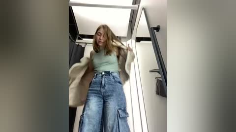 Media: Video of a young woman with light skin and shoulder-length blonde hair, wearing a beige coat over a green top and high-waisted, distressed jeans. She stands in a modern, minimalistic dressing room with a white ceiling, grey walls, and a metal hook.