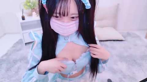 Media: Video of an Asian woman with long black hair in a pastel blue cosplay maid outfit, revealing her large breasts. She wears a pink face mask and kneels on a grey rug in a minimalist room.