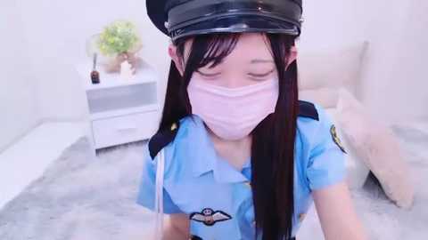 Media: Video of an East Asian woman in a light blue police uniform, wearing a black cap and pink mask, standing on a white carpet in a modern, minimalistic bedroom with a white bed and a white dresser.