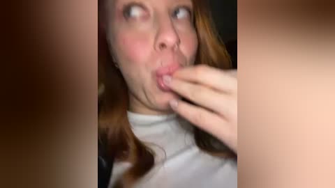 Media: Video of a young woman with fair skin, red hair, and freckles, making a funny face while licking her fingers. She wears a white top. The background is blurred and dark.