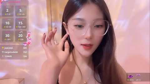 Media: Video of an Asian woman with long black hair, fair skin, and round glasses, speaking on a phone, wearing a black top, in a soft pink room with a digital clock.