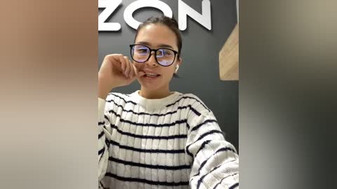 Media: A young woman with dark hair in a ponytail, wearing glasses and a white and black striped sweater, stands in front of a black wall with a \"ZON\" sign, taking a selfie.