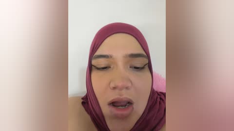 Media: Video of a woman with light brown skin, wearing a maroon hijab, eyes closed, and mouth slightly open, set against a blurred background.