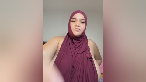 Media: Video of a plus-size woman with medium brown skin, wearing a maroon hijab and sleeveless top, smiling softly. Background is blurred, with soft lighting.