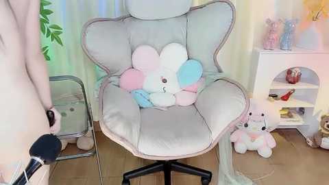 Media: Video of a plush pink cloud pillow with a blue and white bunny plush toy, on a light grey chair. Background features a white shelving unit, green curtains, and a brown floor.