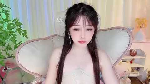 Media: A video of a young Asian woman with long black hair, fair skin, and light makeup, wearing a white strapless dress, sitting in a plush, wingback chair in a softly lit room with greenery and decorative items.