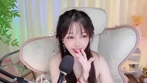 Media: Video of an Asian woman with long dark hair, light skin, wearing a white top, sitting on a white chair, smiling and licking her finger near a microphone.