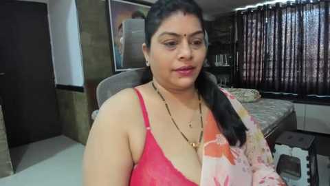 Media: Video of a middle-aged South Asian woman in a pink lace bra, wearing a floral saree, sitting in a dimly-lit, cluttered bedroom with a bed, curtains, and wall art.