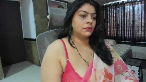 Media: Video of a South Asian woman with medium skin tone, wearing a pink and floral sari, sitting on a chair in a modestly decorated bedroom with brown curtains and a bed.