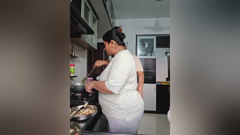 Media: Video of a woman with dark hair tied up in a bun, wearing a white chef's jacket and grey pants, cooking in a modern kitchen with stainless steel appliances, glass cabinets, and a tiled floor.