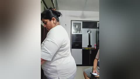 Media: Video of a plus-size woman in a white t-shirt, standing in a modern kitchen with stainless steel appliances and black cabinets.