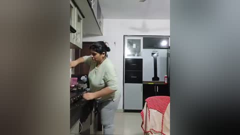 Media: Video of a South Asian woman with medium skin tone and short black hair, cooking in a modern kitchen with white cabinets and stainless steel appliances. She wears a light green shirt and gray pants.