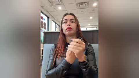 Media: Video of a young woman with long, straight auburn hair, wearing a black leather jacket, seated in an office with fluorescent lighting, holding a pen, looking towards the camera.