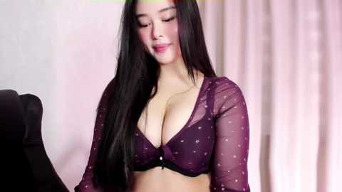 Media: Video of a fair-skinned, East Asian woman with long, straight black hair, wearing a sheer purple bra with star patterns and a matching sheer top, standing in a softly lit, pink-draped room.