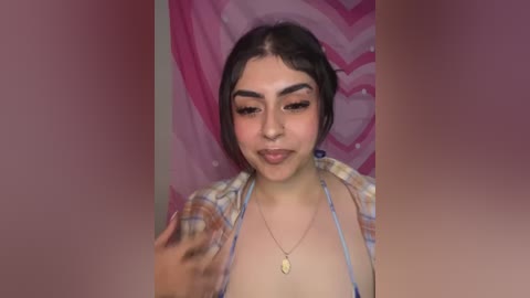 Media: Video of a young Latina woman with medium skin tone, dark hair in a messy bun, wearing a blue bikini top, necklace, and plaid shirt, against a pink heart-patterned backdrop.