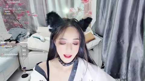 Media: Video of an East Asian woman with long black hair, wearing black cat ears, a black choker, and a white blouse. Background features a white couch, floral decor, and gray curtains.