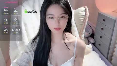 Media: A video of an Asian woman with long black hair, wearing glasses, a white camisole, and sitting on a bed with plush toys, in a bedroom setting.