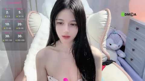 Media: A video of a young, fair-skinned, slim Asian woman with long black hair, wearing a strapless top, sitting on a bed with a plush toy and a white pillow.