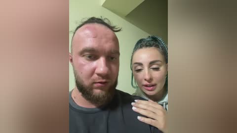 Media: Video of a bald, bearded man with a swollen face, holding a woman with braided hair, wearing a black shirt, in a beige room.