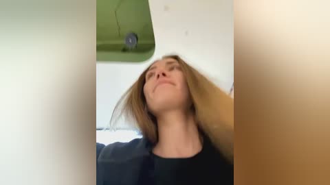 Media: Video of a Caucasian woman with long, light brown hair, wearing a black top, standing in a bright room with green ceiling tiles, looking relaxed and content.