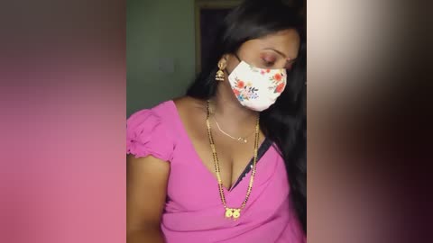 Media: Video of a young South Asian woman with medium brown skin, wearing a pink V-neck blouse, floral face mask, gold necklace, and floral earrings. Background is blurred, featuring a doorway and green wall.