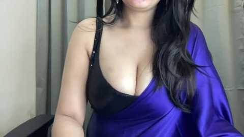 Media: Video of a woman with long, wavy black hair, wearing a deep blue silk sari with a black bra, revealing ample cleavage. Background features light-colored walls and a door.