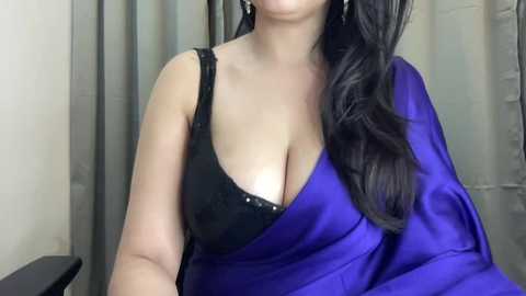 Media: Video of a woman with light brown skin, wearing a black lace bra and a vibrant blue sari, draped elegantly. Her long, wavy black hair cascades over her shoulder. Background features neutral-colored curtains.