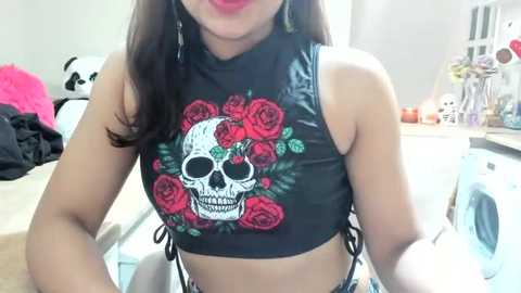 Media: Video of a light-skinned woman with long dark hair, wearing a black sleeveless crop top with a skull and roses design, sitting in a cluttered bedroom with a pink stuffed animal, laundry, and floral decorations.