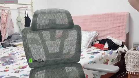 Media: A video of a cluttered bedroom with a gray, mesh office chair, a messy bed covered in colorful bedding, clothes, and a pink blanket.