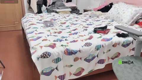 Media: Video of a messy bedroom with a bed covered in colorful fish-patterned bedding, clothes scattered around, and a grey office chair in the foreground.