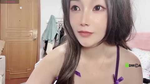 Media: Video of a young East Asian woman with long, dark hair and light skin, wearing a purple bra, in a bedroom with wooden furniture and hanging clothes.