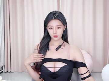 Media: Video of a slender, light-skinned Asian woman with long black hair, wearing a revealing black off-shoulder top with cutouts, sitting indoors against a beige curtain background.