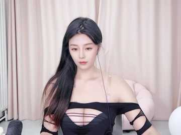 Media: Video of an East Asian woman with long black hair, fair skin, wearing a black off-shoulder top revealing cleavage, sitting against beige curtains, headphones on.