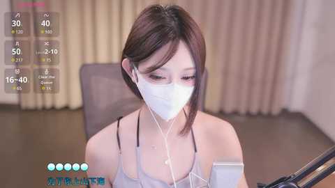 Media: Video of an East Asian woman with short brown hair, wearing a white mask, listening to earbuds, seated in a modern room with beige curtains.