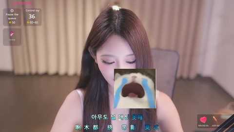 Media: A young Asian woman with long brown hair, wearing a white top, sits in a room with beige curtains. Her face is obscured by a GIF of a crying dog.