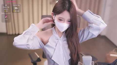 Media: Video of an Asian woman with long brown hair, wearing a white face mask, blue shirt, adjusting headphones, seated at a desk, in a modern office.