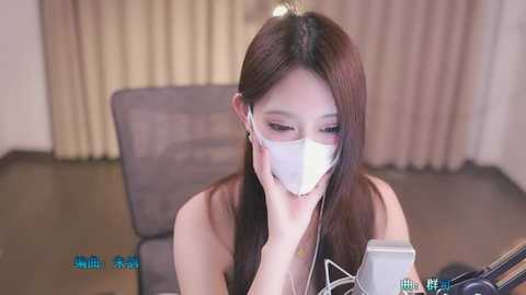 Media: Video of a young East Asian woman with long brown hair, wearing a white face mask, listening to white earbuds, seated in a beige room with a gray couch and light brown curtains.