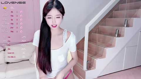 Media: Video of a slender, light-skinned East Asian woman with long black hair, wearing a white top and pink shorts, standing on a beige carpeted staircase in a modern, light-colored room.