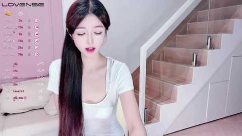 Media: Video of a young East Asian woman with long, straight black hair, wearing a white top, sitting on a white couch. Background features a pink wall calendar and wooden stairs.