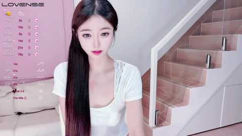 Media: Video of an Asian woman with long black hair, light skin, and red lipstick, wearing a white top, standing indoors by a staircase with beige walls and a calendar.