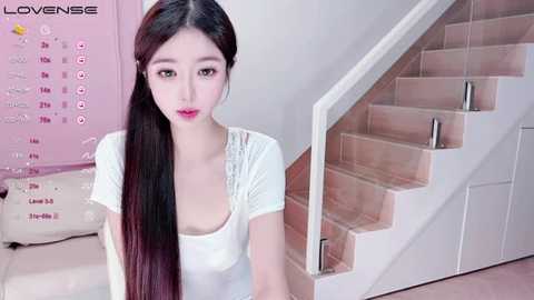 Media: Video of a young Asian woman with long black hair, wearing a white top, sitting on stairs in a pastel-colored room.