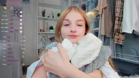 Media: A video of a young Caucasian woman with blonde hair, holding a gray and white plush toy, in a modern, minimalist room with white shelves and hanging clothes.