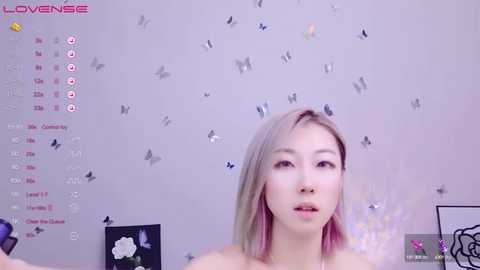 Media: Video of a fair-skinned, young Asian woman with straight, shoulder-length blonde hair, wearing a black top, standing against a pastel pink wall adorned with butterflies and a calendar. Background includes a pink flower and a butterfly figurine.