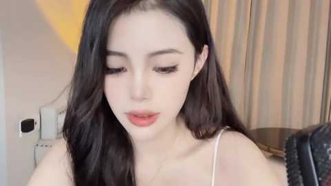 Media: A video of an East Asian woman with fair skin, long black hair, and minimal makeup. She wears a white spaghetti-strap top, has a calm expression, and is indoors with beige curtains and a black couch in the background.