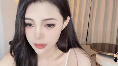 Media: Video of an East Asian woman with fair skin, long black hair, and dark eyes. She wears a white top and sits indoors near beige curtains and a round mirror.