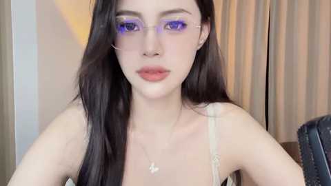 Media: Video of a young woman with pale skin, long black hair, purple eyeshadow, and glasses, wearing a white camisole and silver necklace. She stands against beige curtains in a dimly lit room.