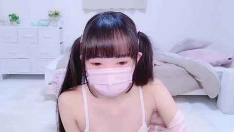 Media: Video of an Asian woman with long black hair in pigtails, wearing a pink mask and light pink spaghetti strap top, sitting on a white bed in a minimalist, white room.