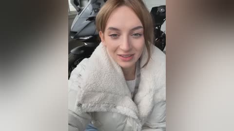 Media: Video of a young woman with fair skin, light brown hair, and blue eyes, wearing a white fleece jacket, smiling while sitting in the driver's seat of a car.