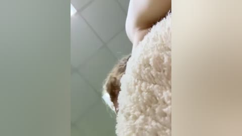 Media: Video of a woman's bare stomach with a small, light-colored mole, wearing a cream-colored, fuzzy sweater, in a tiled room with a white ceiling.