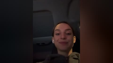 Media: Video of a young woman with fair skin, dark hair, and light makeup, smiling in a car's rearview mirror, wearing a green shirt, dimly lit.